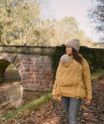 A woman wearing the Mamalila Allweather Cosy Allrounder Babywearing Jacket in a Mustard yellow colour carrying a baby using the babywearing insert.