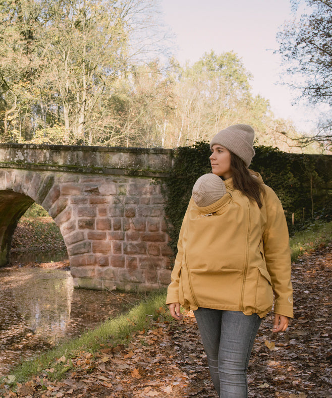 A woman wearing the Mamalila Allweather Cosy Allrounder Babywearing Jacket in a Mustard yellow colour carrying a baby using the babywearing insert.