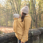 A woman wearing the Mamalila Allweather Cosy Allrounder Babywearing Jacket in a Mustard yellow colour carrying a baby using the babywearing insert.