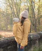 A woman wearing the Mamalila Allweather Cosy Allrounder Babywearing Jacket in a Mustard yellow colour carrying a baby using the babywearing insert.