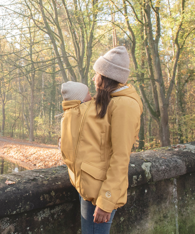 A woman wearing the Mamalila Allweather Cosy Allrounder Babywearing Jacket in a Mustard yellow colour carrying a baby using the babywearing insert.
