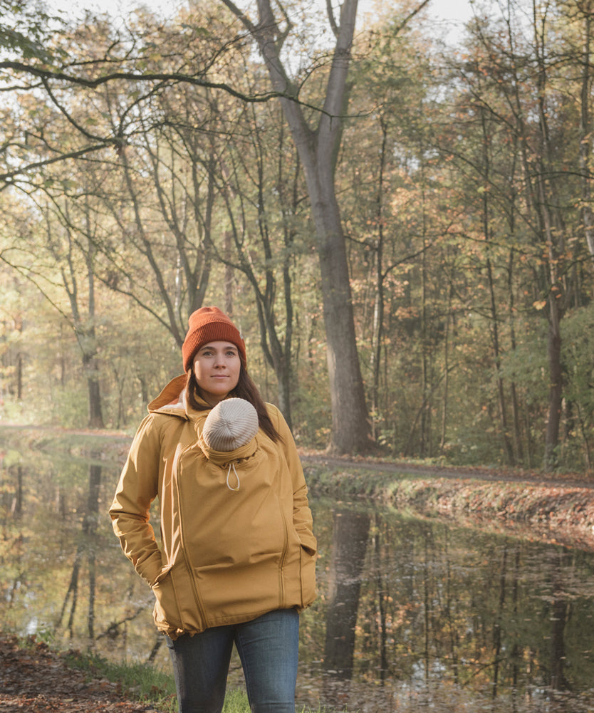 A woman wearing the Mamalila Allweather Cosy Allrounder Babywearing Jacket in a Mustard yellow colour carrying a baby using the babywearing insert.