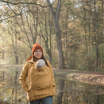 A woman wearing the Mamalila Allweather Cosy Allrounder Babywearing Jacket in a Mustard yellow colour carrying a baby using the babywearing insert.