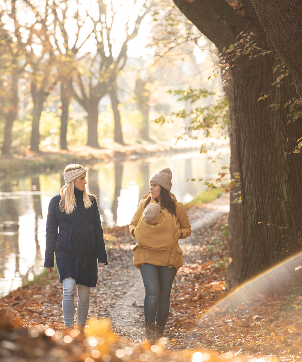 Two women walking on a path surrounded by fallen leaves. One is wearing a Mamalila Eco Wool Oslo Babywearing Coat and the other is wearing a Mamalila Allweather Cosy Allrounder Mustard Babywearing Jacket.