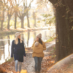 Two women walking on a path surrounded by fallen leaves. One is wearing a Mamalila Eco Wool Oslo Babywearing Coat and the other is wearing a Mamalila Allweather Cosy Allrounder Mustard Babywearing Jacket.