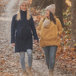 Two women walking on a path surrounded by fallen leaves. One is wearing a Mamalila Eco Wool Oslo Babywearing Coat and the other is wearing a Mamalila Allweather Cosy Allrounder Mustard Babywearing Jacket.