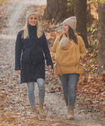 Two women walking on a path surrounded by fallen leaves. One is wearing a Mamalila Eco Wool Oslo Babywearing Coat and the other is wearing a Mamalila Allweather Cosy Allrounder Mustard Babywearing Jacket.