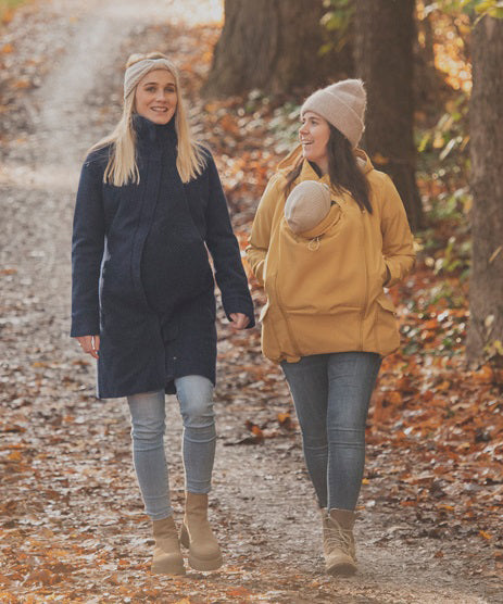 Two women walking on a path surrounded by fallen leaves. One is wearing a Mamalila Eco Wool Oslo Babywearing Coat and the other is wearing a Mamalila Allweather Cosy Allrounder Mustard Babywearing Jacket.
