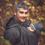 Mamalila Shelter Men's Babywearing Rain Jacket in Anthracite. Lifestyle view of this men's technical babywearing rain coat with the baby carrier insert on the front. Woodland background 