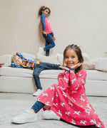 A child sitting on a floor wearing the Maxomorra Unicorn Long Sleeve Circle Dress. There are two children seen in the background  on a sofa also wearing Maxomorra. 
