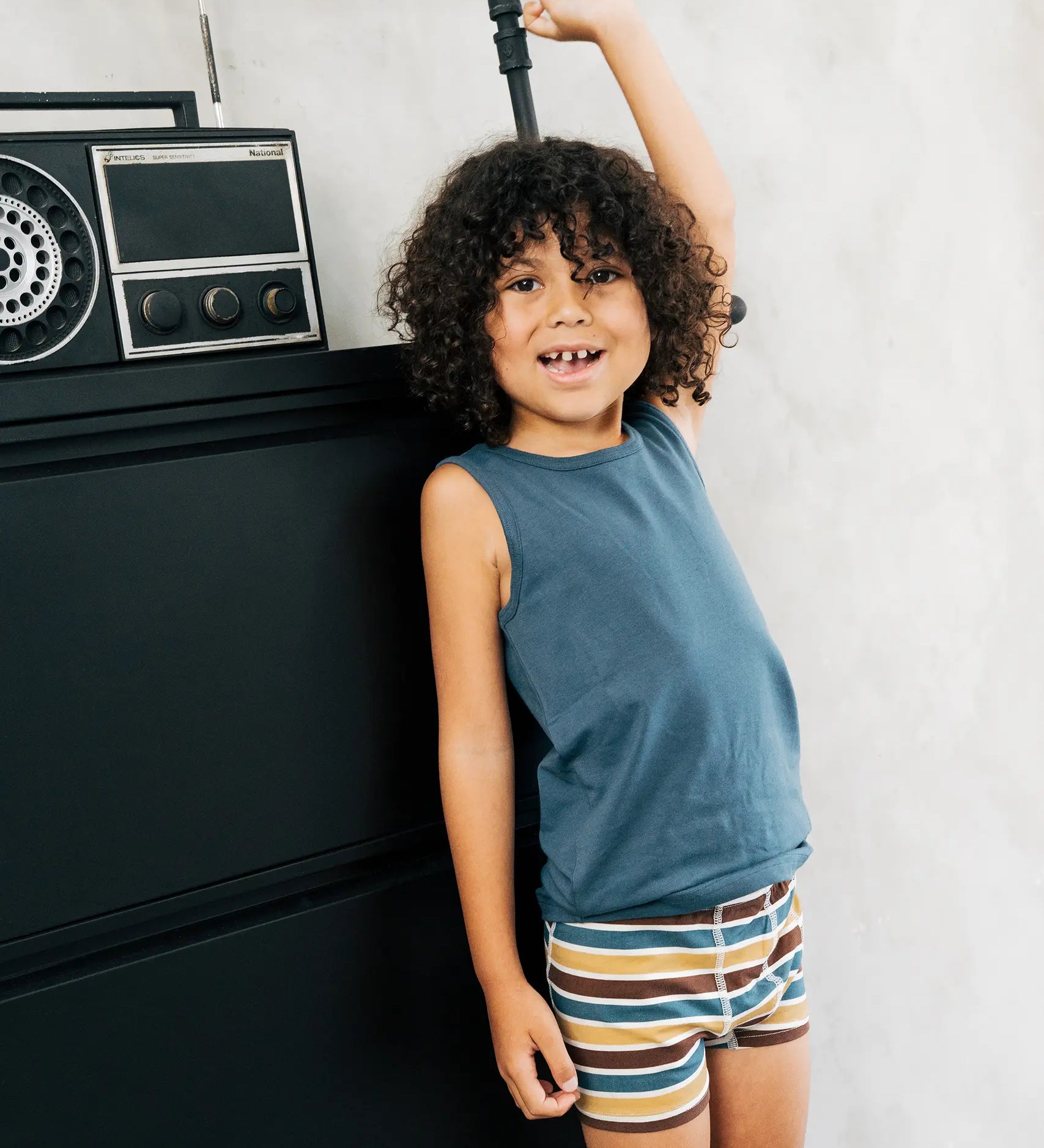 Child wearing the Maxomorra kids GOTS organic cotton boxer shorts in a blue, white, yellow and brown striped design with a blue vest top.