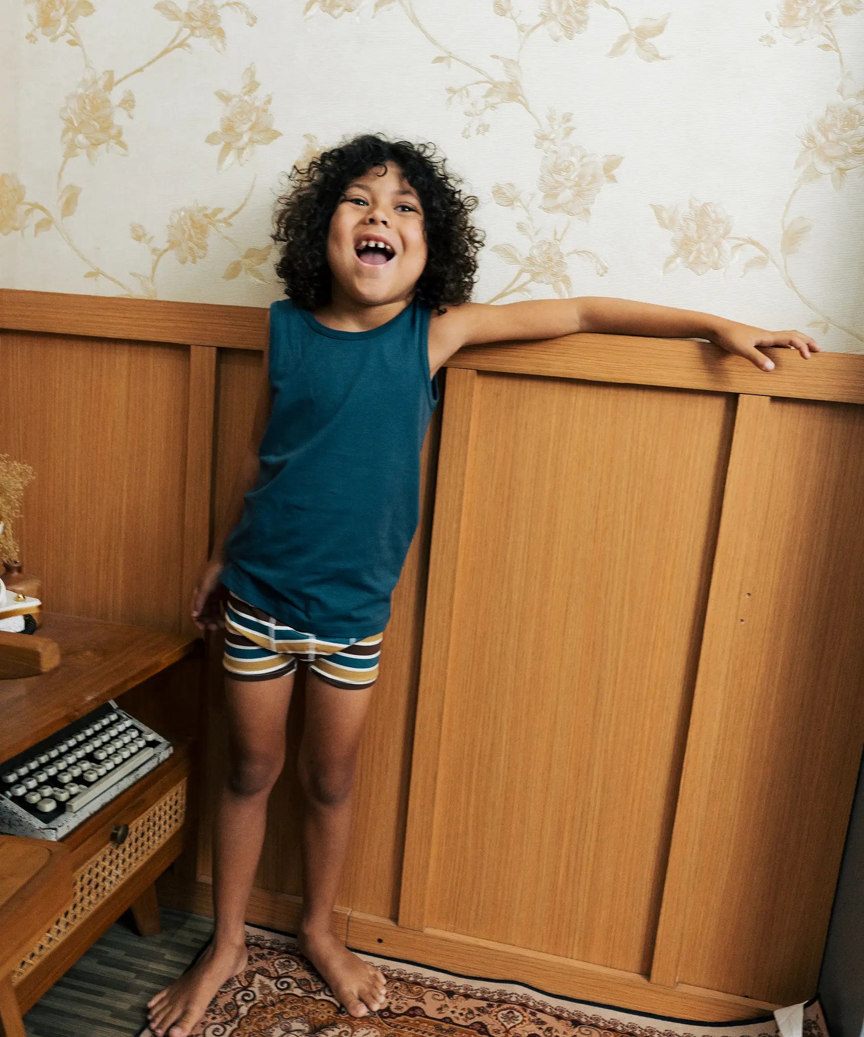 Child wearing the Maxomorra kids GOTS organic cotton boxer shorts in a blue, white, yellow and brown striped design with a blue vest top.