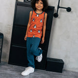 Child wearing the Maxomorra kids dragons organic cotton sleeveless vest with blue teal joggers. Rust red sleeveless vest top with repeat print featuring a blue dragon and yellow fire outlines with blue binding on the neck and arm holes. 
