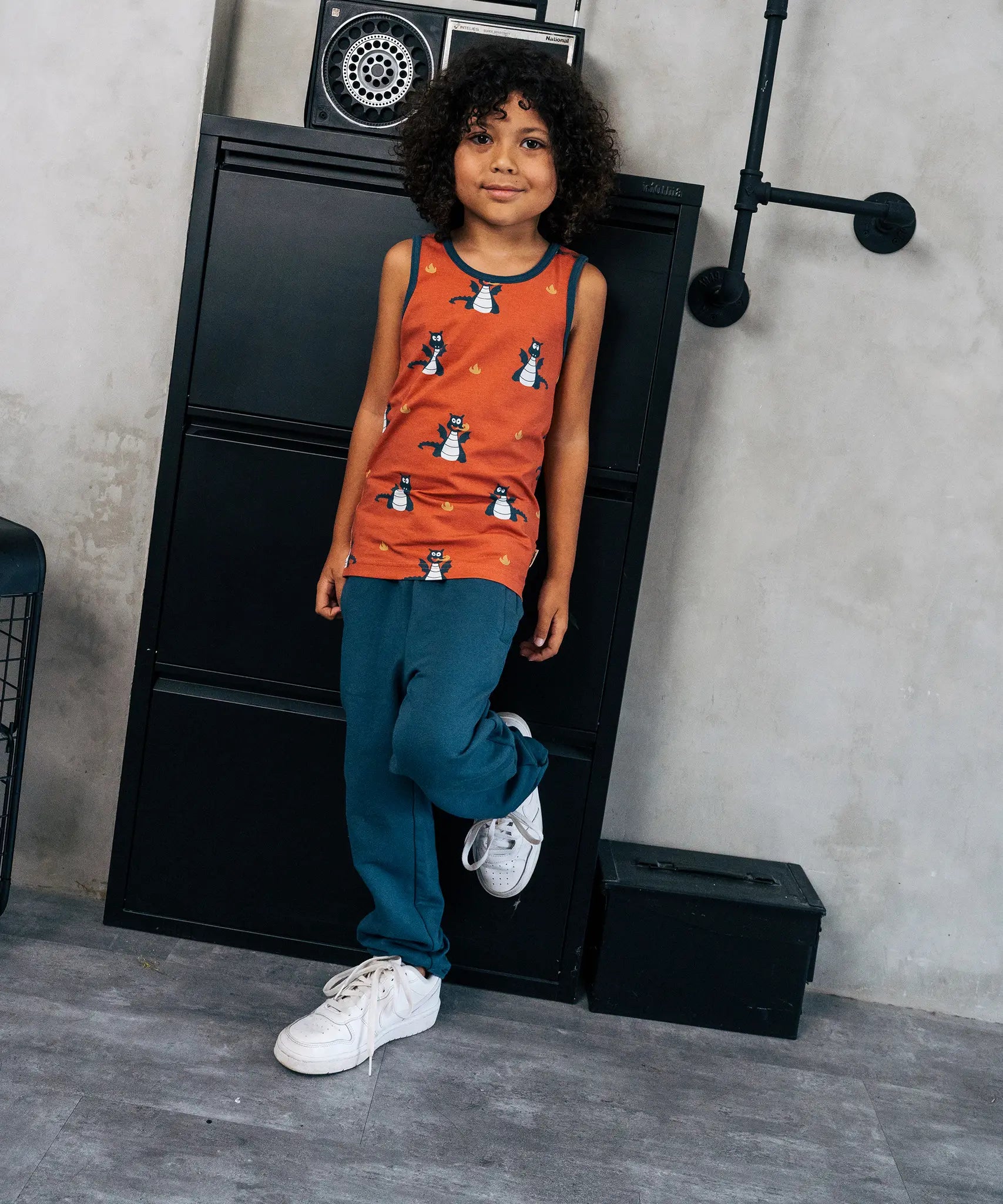 Child wearing the Maxomorra kids dragons organic cotton sleeveless vest with blue teal joggers. Rust red sleeveless vest top with repeat print featuring a blue dragon and yellow fire outlines with blue binding on the neck and arm holes. 
