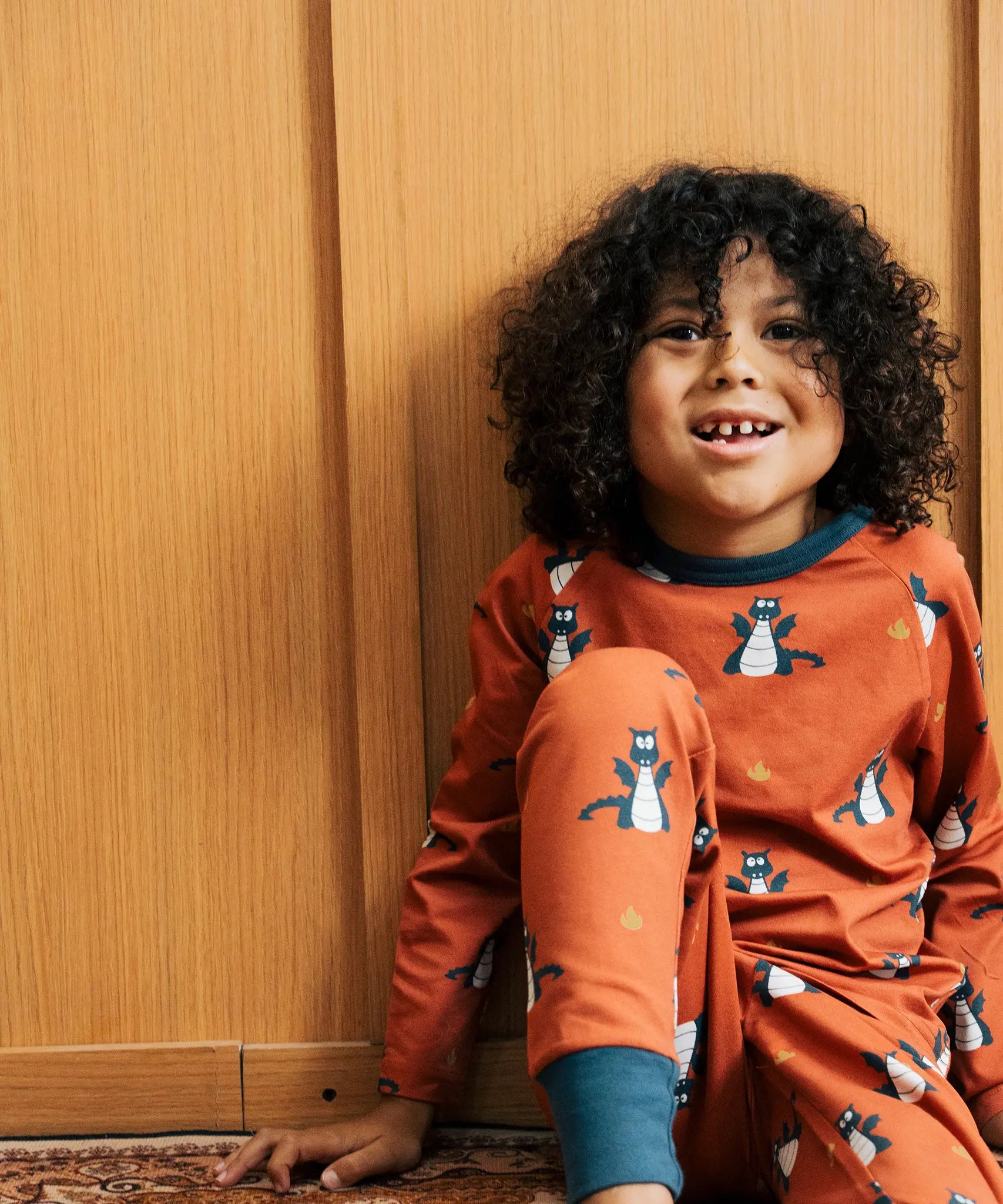 Child wearing the Maxomorra kids dragons organic cotton long sleeve top with pyjama trousers. Red top with repeat blue dragon print and blue arm cuffs and binding on the neck. 
