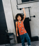Child wearing the Maxomorra kids dragons organic cotton sleeveless vest with teal joggers. Rust red sleeveless vest top with repeat print featuring a blue dragon and yellow fire outlines with blue binding on the neck and arm holes. 
