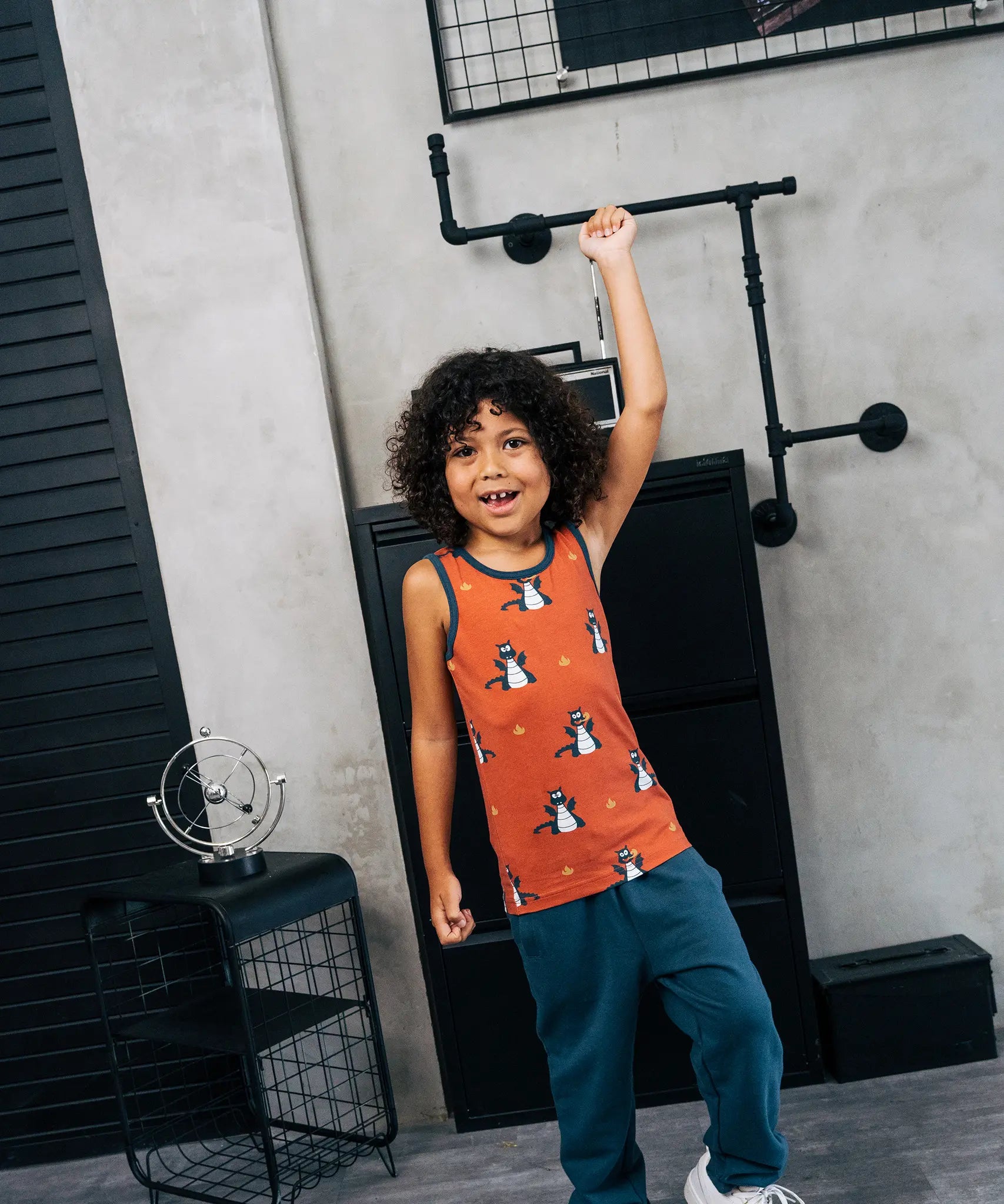 Child wearing the Maxomorra kids dragons organic cotton sleeveless vest with teal joggers. Rust red sleeveless vest top with repeat print featuring a blue dragon and yellow fire outlines with blue binding on the neck and arm holes. 
