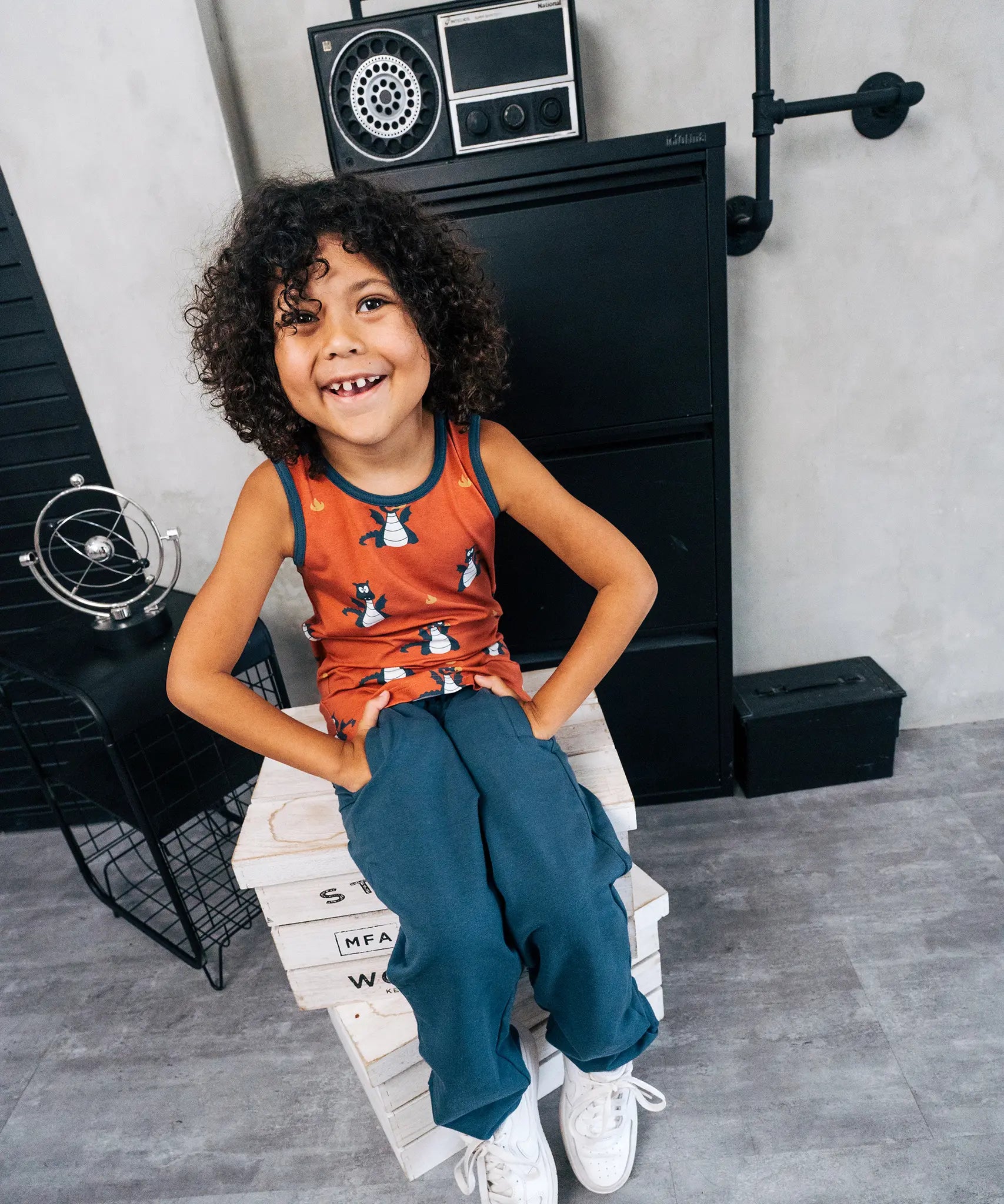 Child wearing the Maxomorra kids dragons organic cotton sleeveless vest with blue teal joggers. Rust red sleeveless vest top with repeat print featuring a blue dragon and yellow fire outlines with blue binding on the neck and arm holes. 
