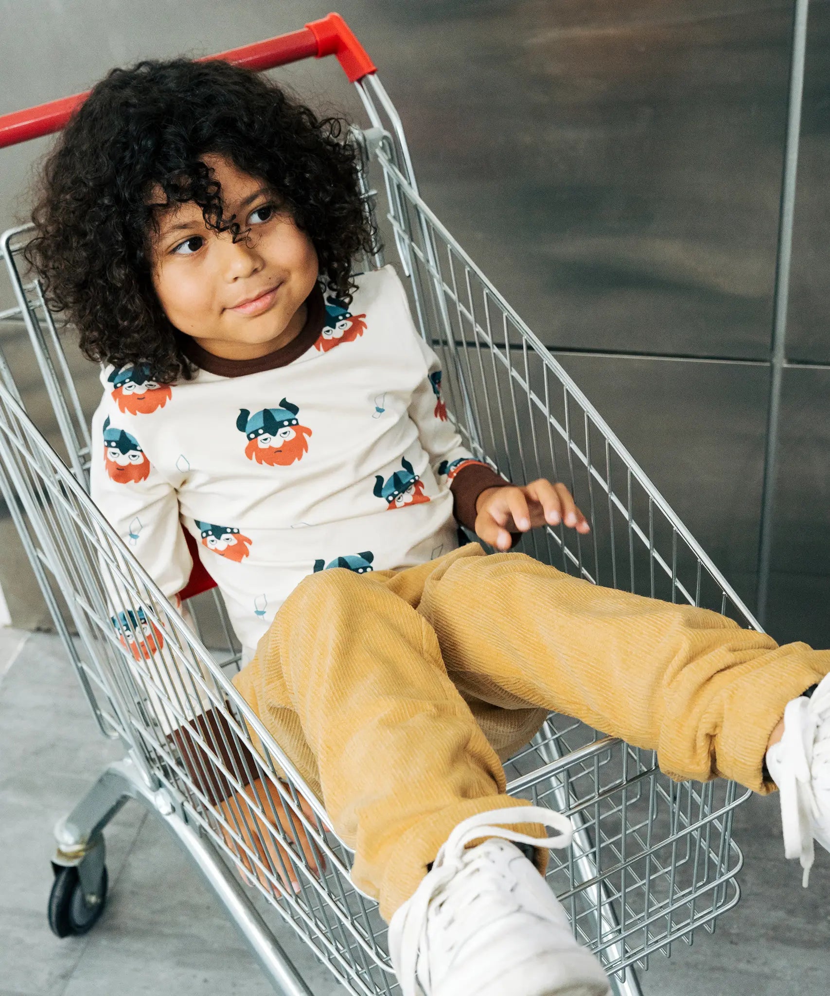 Child wearing the Maxomorra kids vikings organic cotton long sleeve top with mustard yellow trousers. Light coloured top with  repeat viking print and contrasting brown arm cuffs and binding on the neck. 
