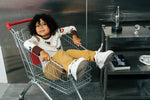 Child wearing the Maxomorra kids vikings organic cotton long sleeve top with mustard yellow trousers siting in a trolley. Light coloured top with  repeat viking print and contrasting brown arm cuffs and binding on the neck. 
