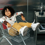 Child wearing the Maxomorra kids vikings organic cotton long sleeve top with mustard yellow trousers siting in a trolley. Light coloured top with  repeat viking print and contrasting brown arm cuffs and binding on the neck. 
