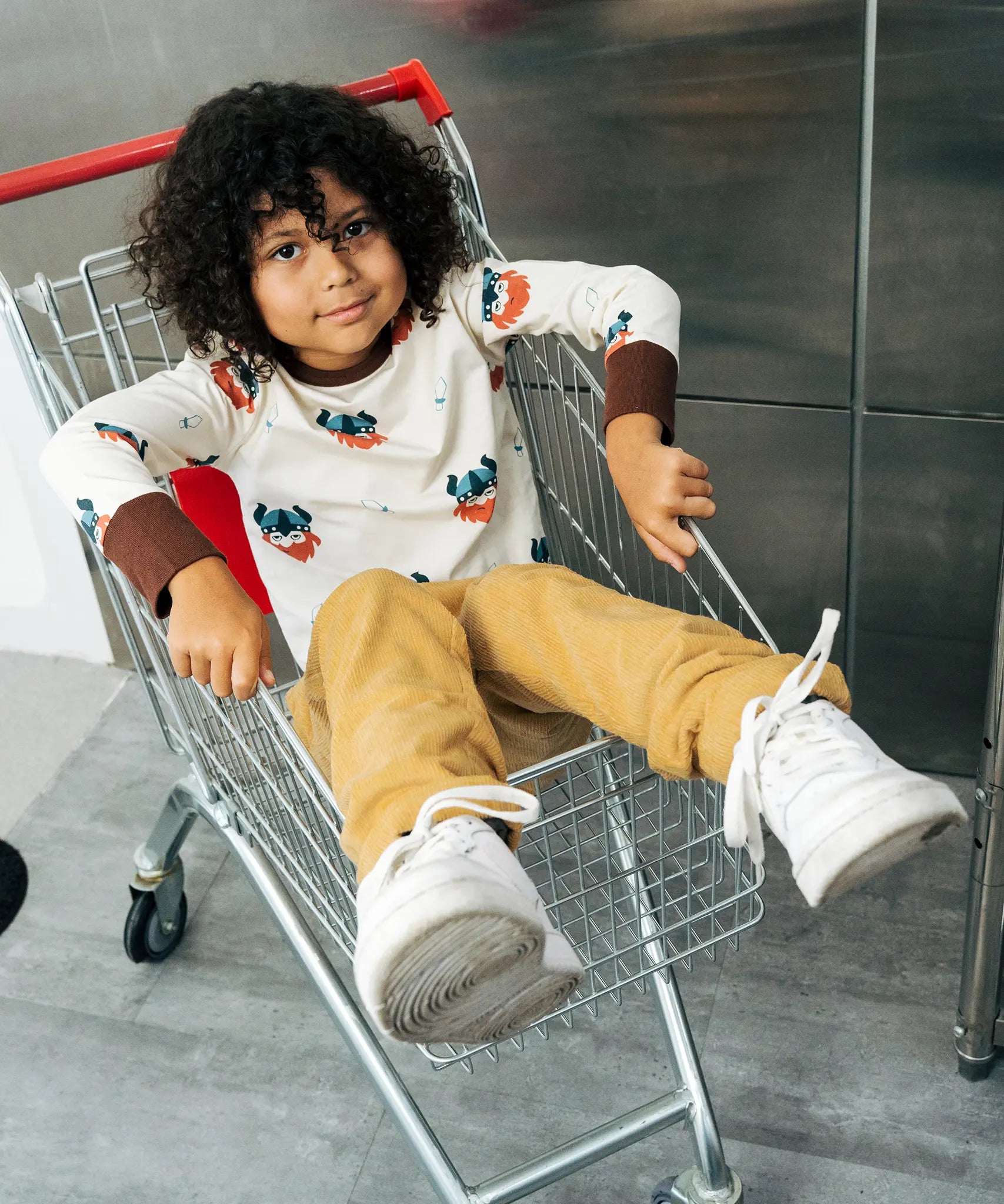 Child wearing the Maxomorra kids vikings organic cotton long sleeve top with mustard yellow trousers. Light coloured top with  repeat viking print and contrasting brown arm cuffs and binding on the neck. 
