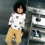 Child wearing the Maxomorra kids vikings organic cotton long sleeve top with mustard yellow trousers. Light coloured top with  repeat viking print and contrasting brown arm cuffs and binding on the neck. 
