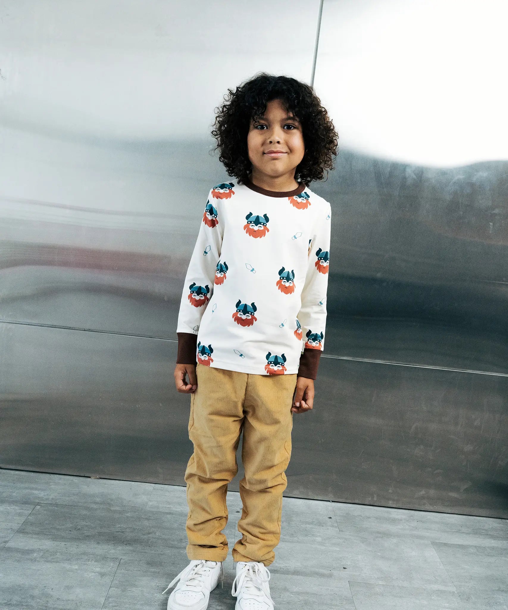 Child wearing the Maxomorra kids vikings organic cotton long sleeve top with mustard yellow trousers. Light coloured top with  repeat viking print and contrasting brown arm cuffs and binding on the neck. 

