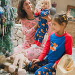 A young child wearing the Maxomorra Reindeer Long Sleeve Rompersuit being held by a woman wearing pink pyjamas. Another child sits in front of them in reindeer print leggings and a matching reindeer raglan top. 
