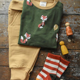 Maxomorra green long sleeved fox print top placed next to mustard yellow trousers and Little Green Radical organic cotton socks on a wooden surface with Grapat peg dolls. 