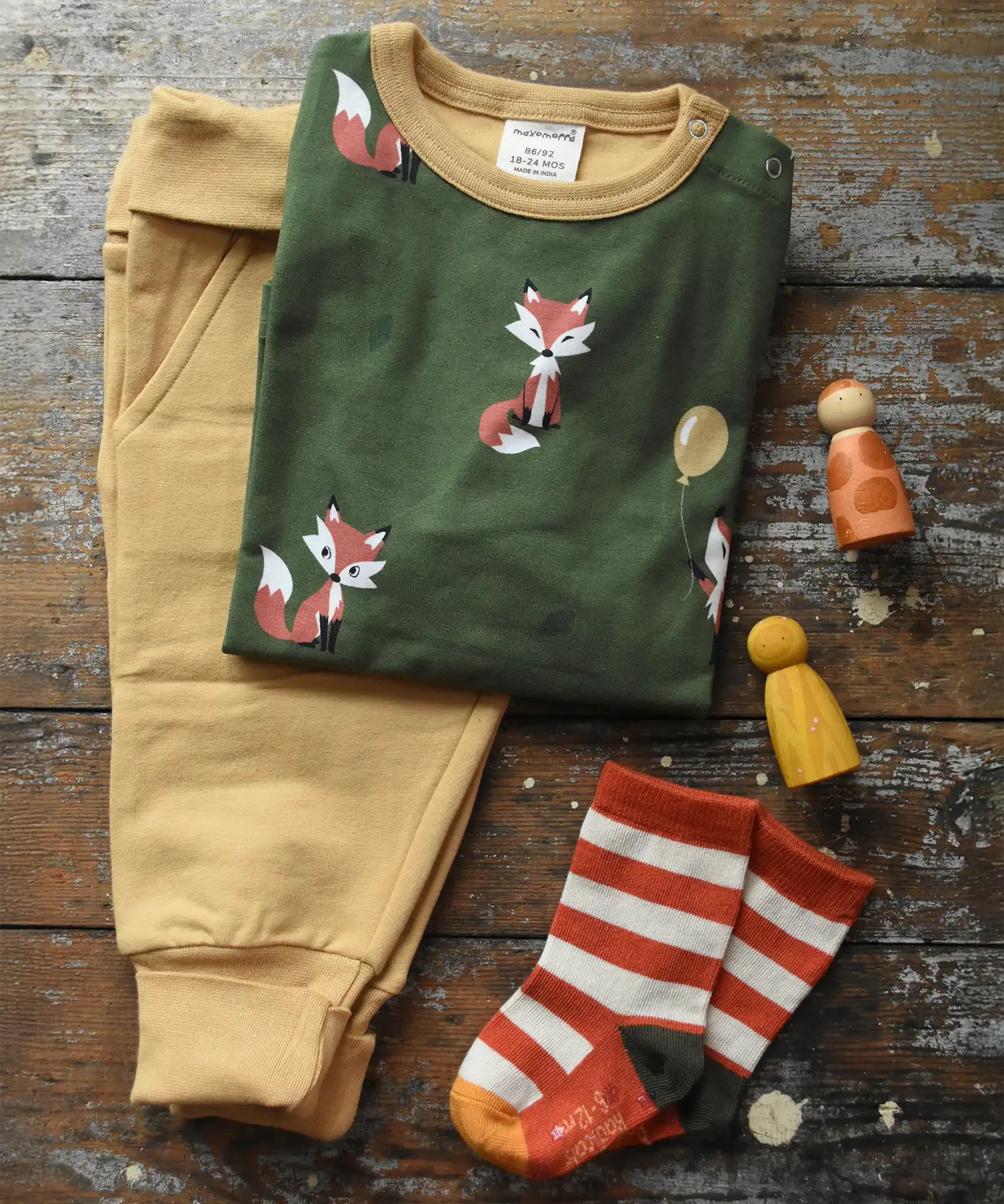 Maxomorra green long sleeved fox print top placed next to mustard yellow trousers and Little Green Radical organic cotton socks on a wooden surface with Grapat peg dolls. 