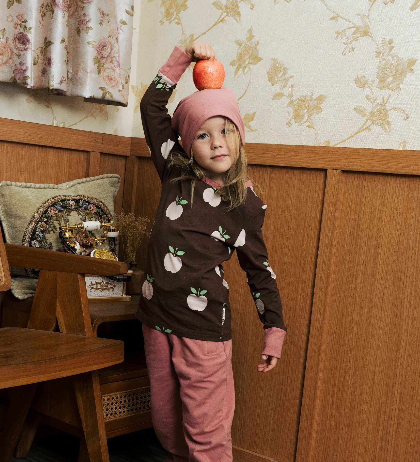 Child wearing the Maxomorra GOTS organic cotton long sleeve top in brown with a cute repeating pink apple print and pale pink neck line and cuffs with pink trousers and beanie style hat.