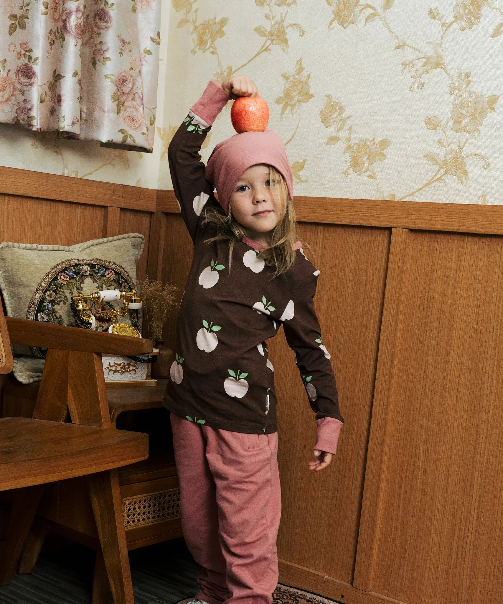 Child wearing the Maxomorra GOTS organic cotton long sleeve top in brown with a cute repeating pink apple print and pale pink neck line and cuffs with pink trousers and beanie style hat.