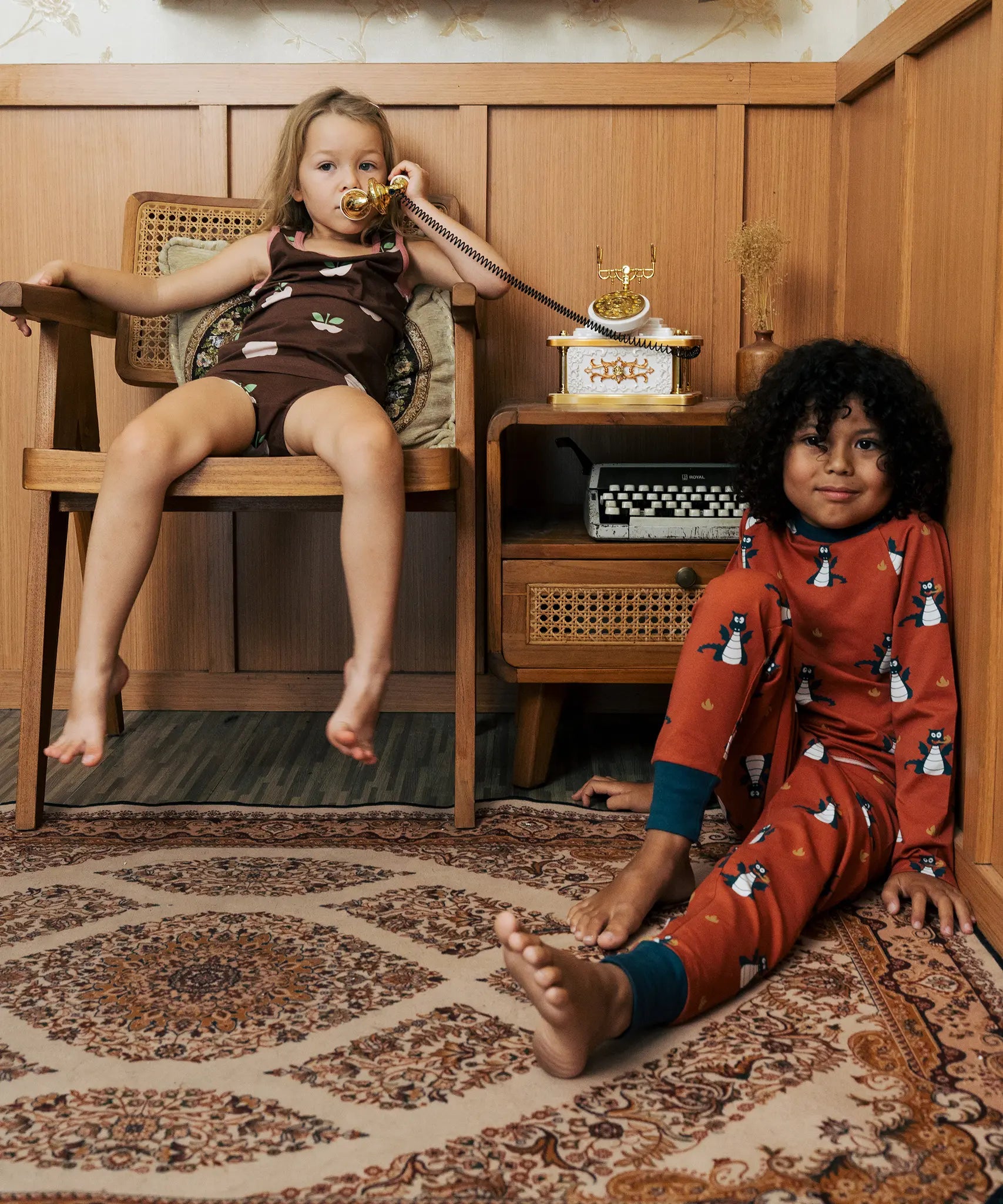 Two Children wearing Maxomorra organic cotton clothing. One is wearing a brown apple print vest and shorts the other is wearing a rust red blue dragon print long sleeved top with matching trousers.