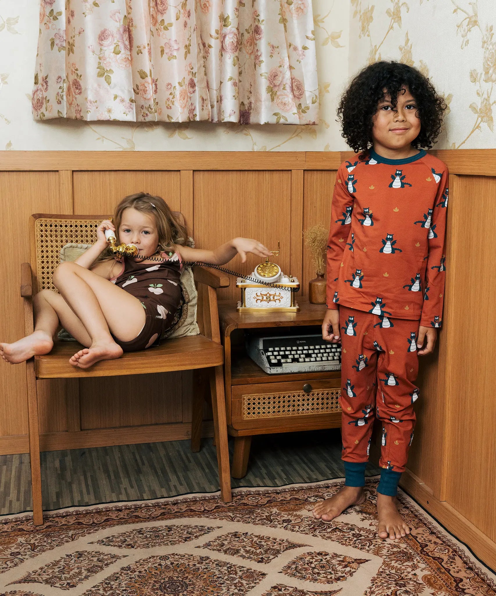 Two Children wearing Maxomorra organic cotton clothing. One is wearing a brown apple print vest and shorts the other is wearing a rust red blue dragon print long sleeved top with matching trousers.