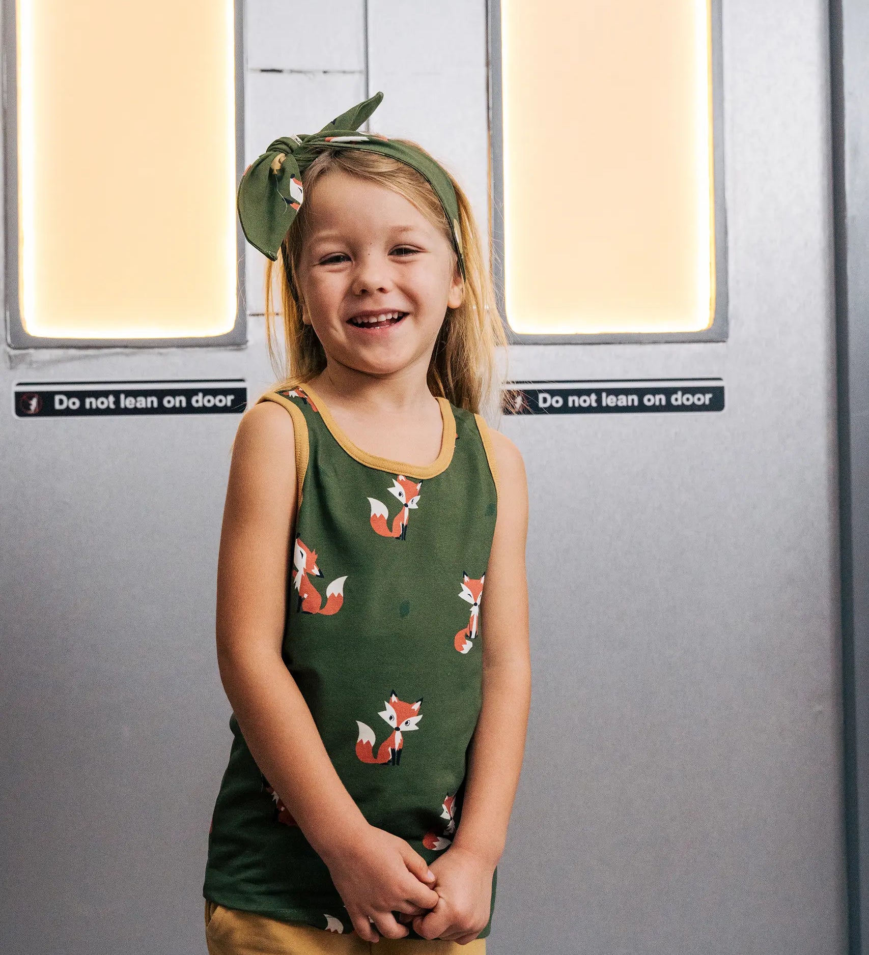 Child wearing the Maxomorra kids organic cotton green fox print vest with yellow trousers on a train.
