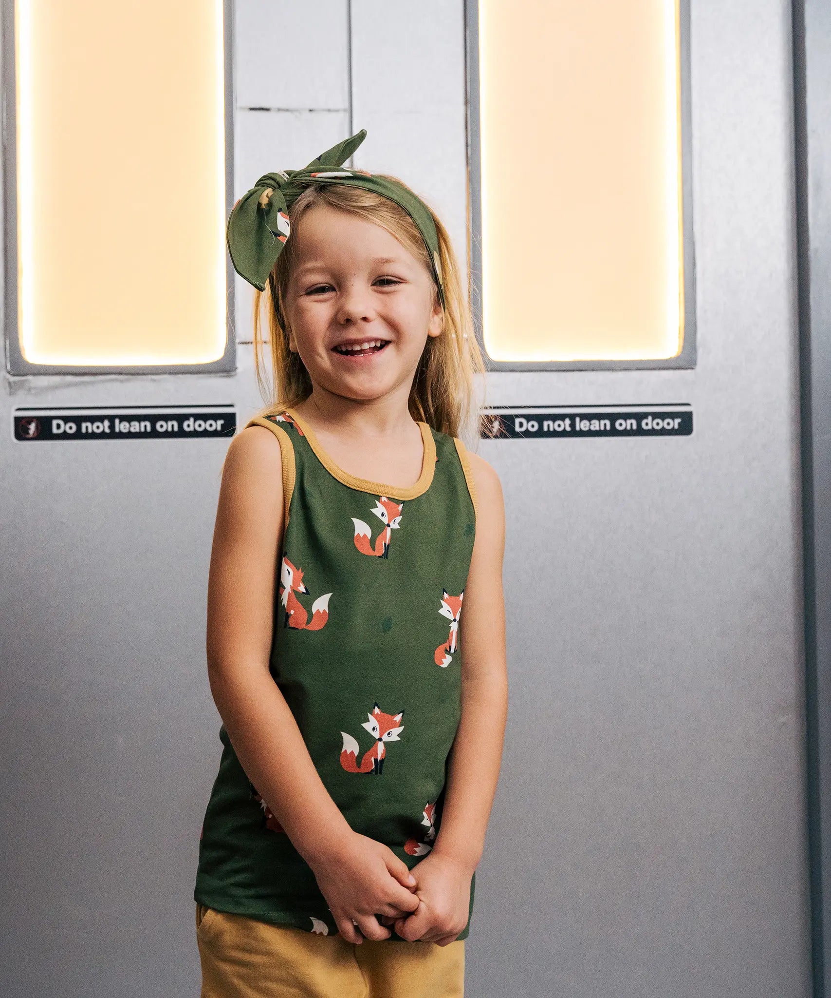 Child wearing the Maxomorra kids organic cotton green fox print vest with yellow trousers on a train.