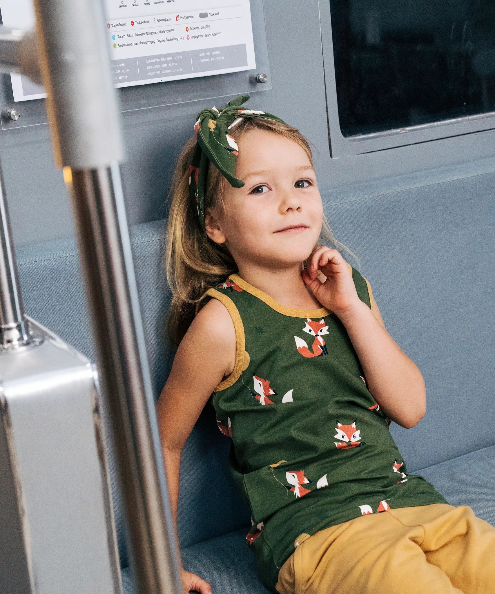 Child wearing the Maxomorra kids organic cotton green fox print vest with yellow trousers sitting on a train.