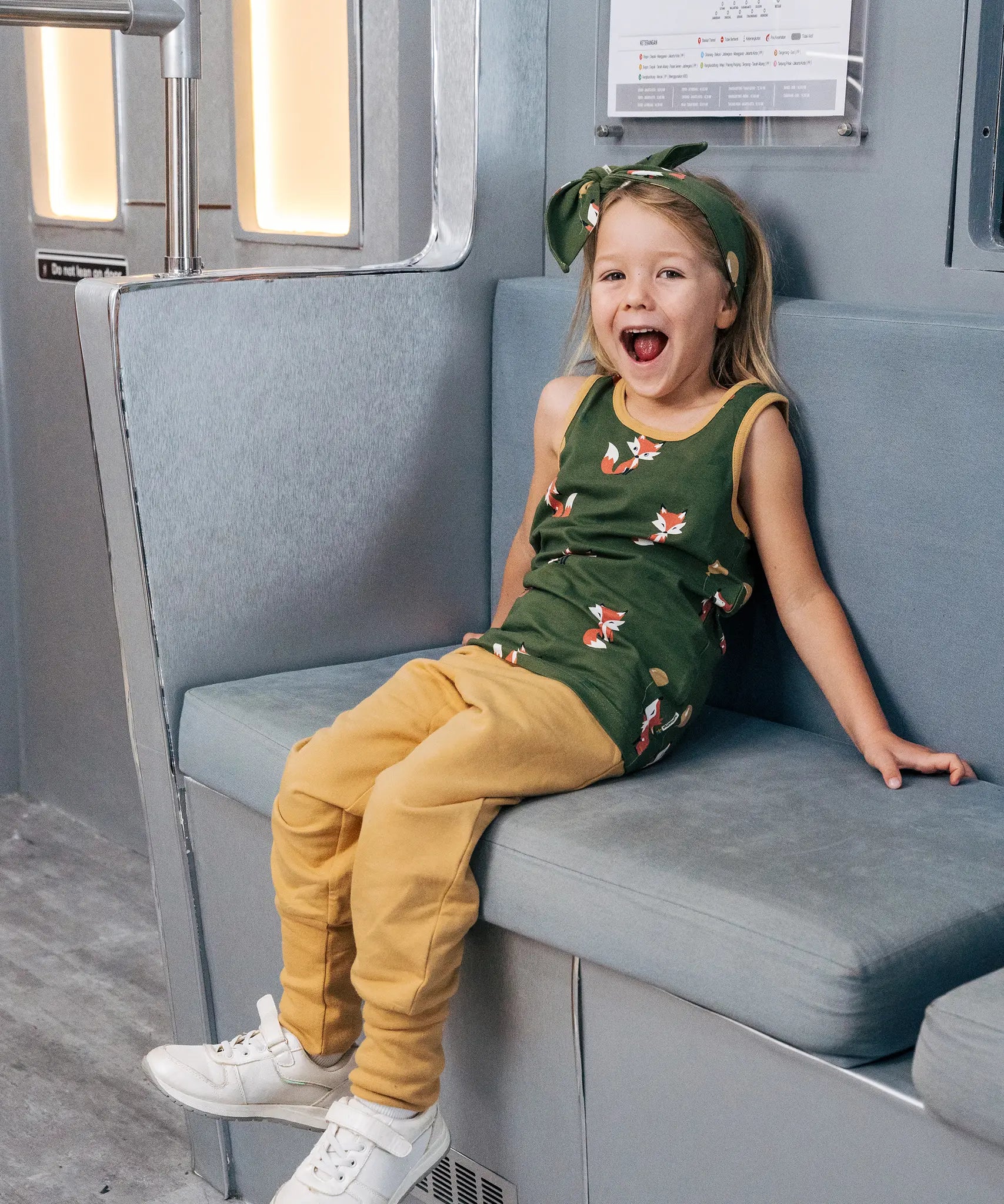 Child wearing the Maxomorra kids organic cotton green fox print vest with yellow trousers sitting on a train.