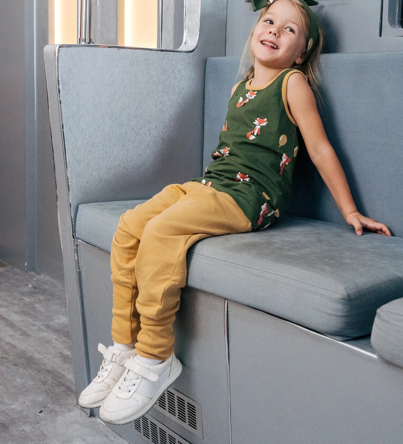 Child wearing the Maxomorra kids organic cotton green fox print vest with yellow trousers sitting on a train.