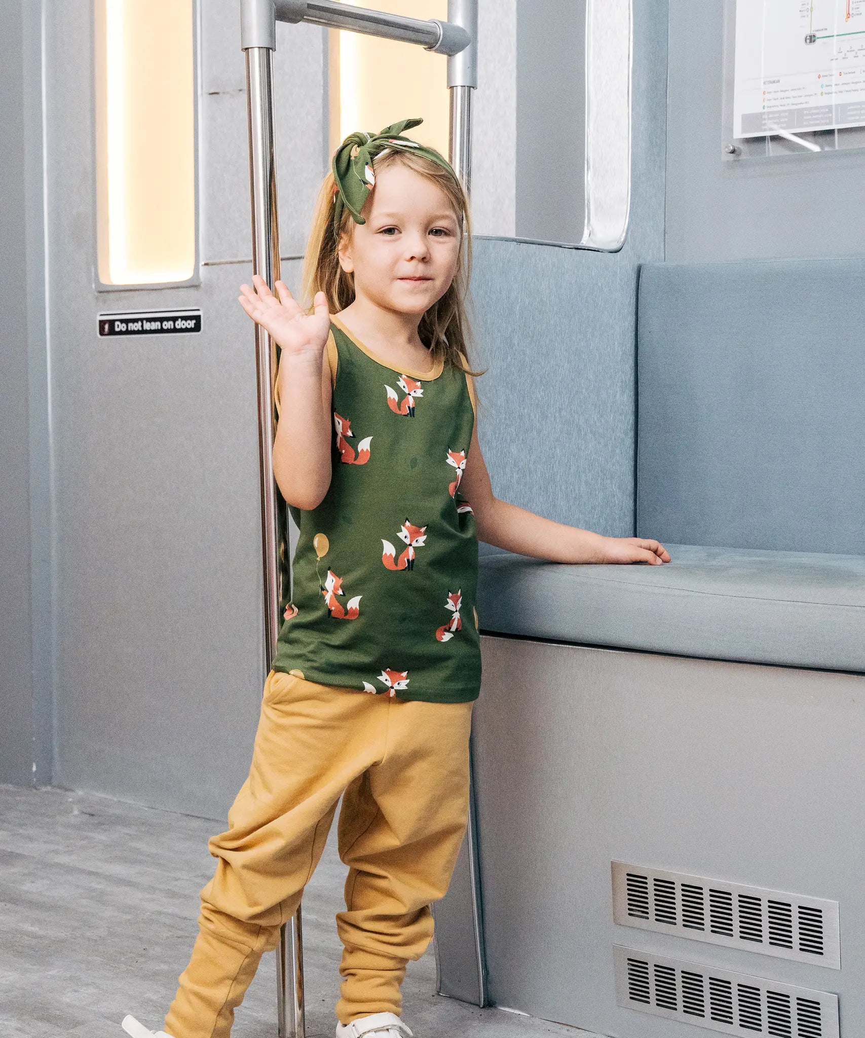 Child wearing the Maxomorra kids organic cotton green fox print vest with yellow trousers standing on a train.