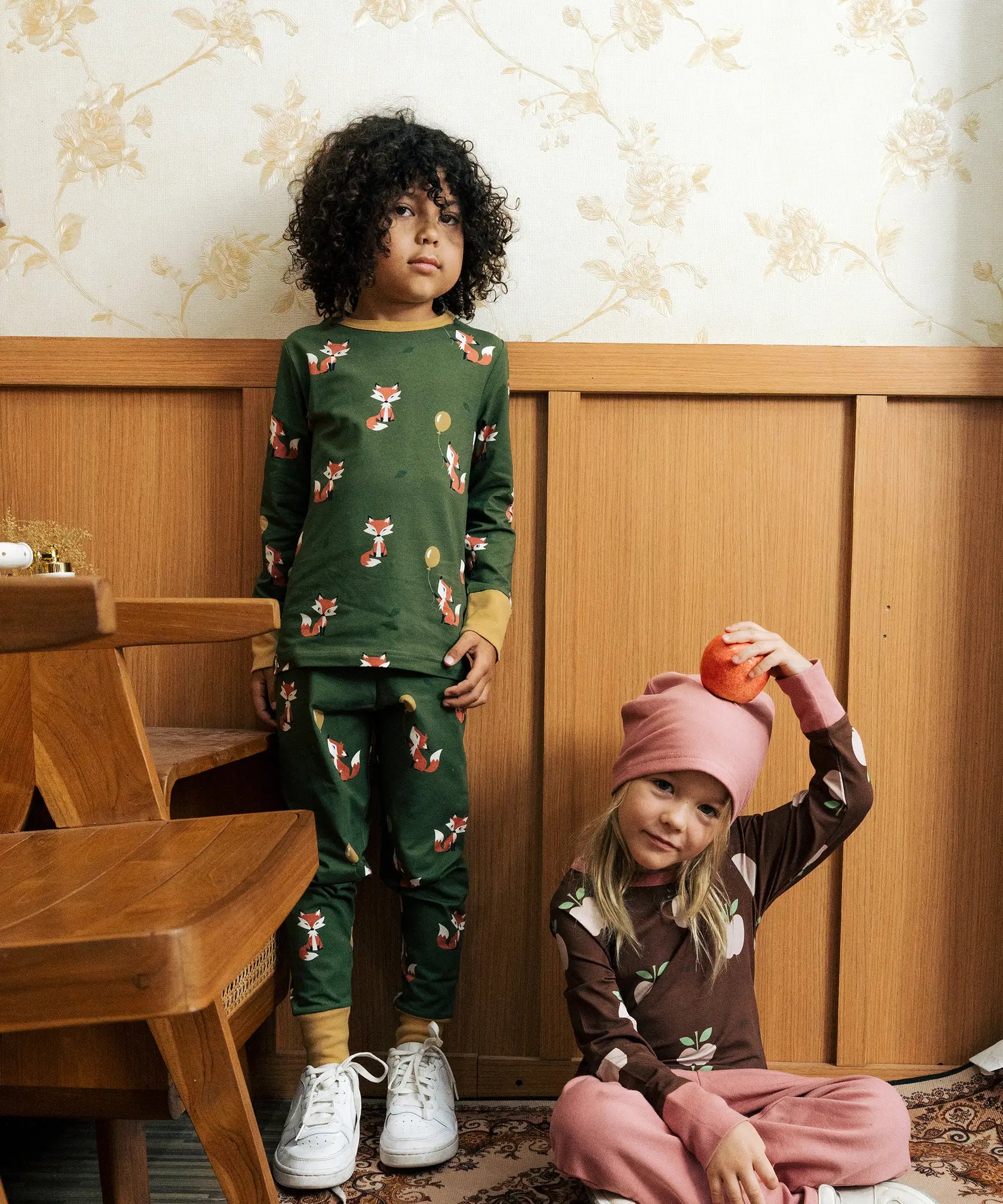Child wearing Maxomora organic cotton green long-sleeve top with all over red fox print with matching pyjama trousers. Another child in background wearing brown apple print top with pink trousers. 