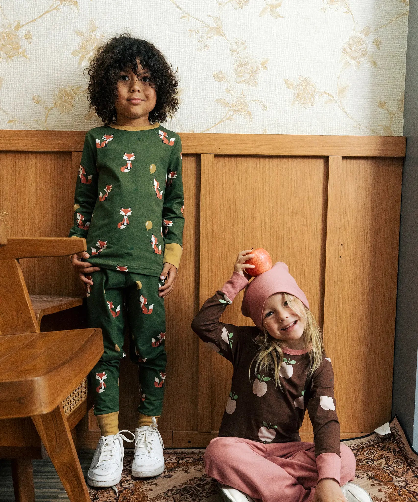 Child wearing Maxomora organic cotton green long-sleeve top with all over red fox print with matching pyjama trousers with a child sitting besides him wearing brown apple print top with pink trousers.