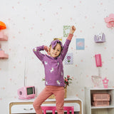 Child wearing the Maxomorra kids unicorns organic cotton long sleeve top with pink trousers. Purple top with  repeat unicorn print and light pink arm cuffs and binding on the neck. 
