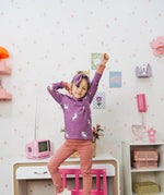 Child wearing the Maxomorra kids unicorns organic cotton long sleeve top with pink trousers. Purple top with  repeat unicorn print and light pink arm cuffs and binding on the neck. 
