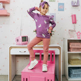 Child wearing the Maxomorra kids unicorns organic cotton long sleeve top with pink leggings. Purple top with  repeat unicorn print and light pink arm cuffs and binding on the neck. 