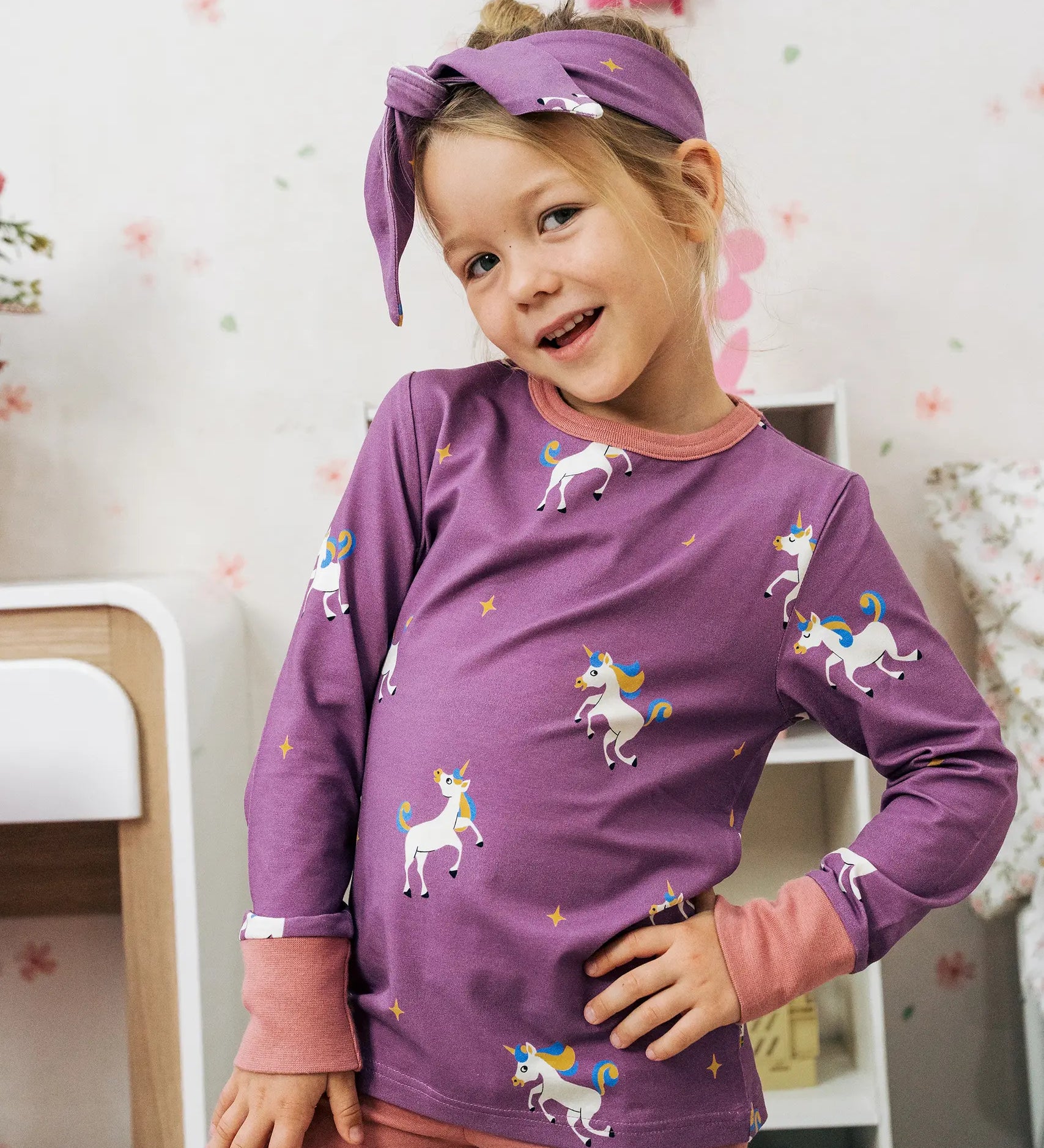 Child wearing the Maxomorra kids unicorns organic cotton long sleeve top. Purple top with  repeat unicorn print and light pink arm cuffs and binding on the neck. 

