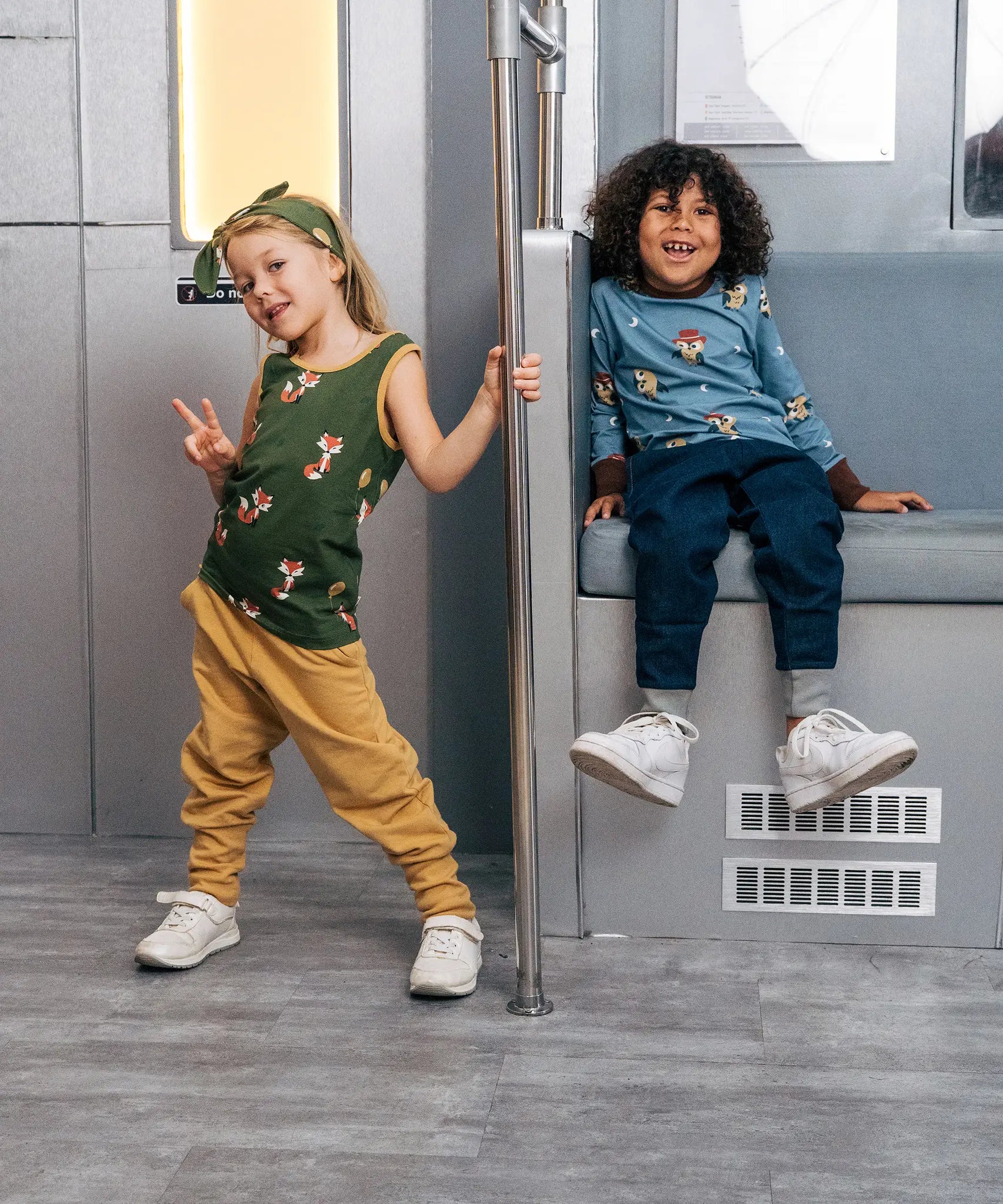 Child wearing the Maxomorra kids organic cotton green fox print vest with yellow trousers on a train with a child wearing a blue owl print long sleeved top and blue trousers sitting next to her .