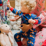 A young child wearing the Maxomorra Reindeer Long Sleeve Rompersuit. The child is reaching for a bauble from the Christmas tree. 