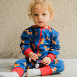 A young child wearing the Maxomorra Reindeer Long Sleeve Rompersuit. The child is sitting in front of a beige coloured wall. 